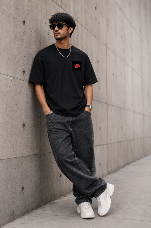 Man wearing a black t-shirt with a red logo, dark jeans, and white sneakers against a concrete wall.